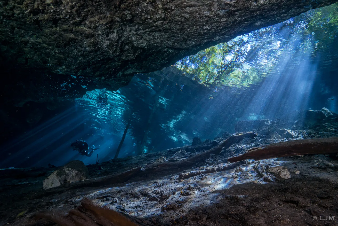 Sunlight streams through a crystal-clear cenote, illuminating rocky formations and clear water, illustrating the deceptive beauty of cavern dives.