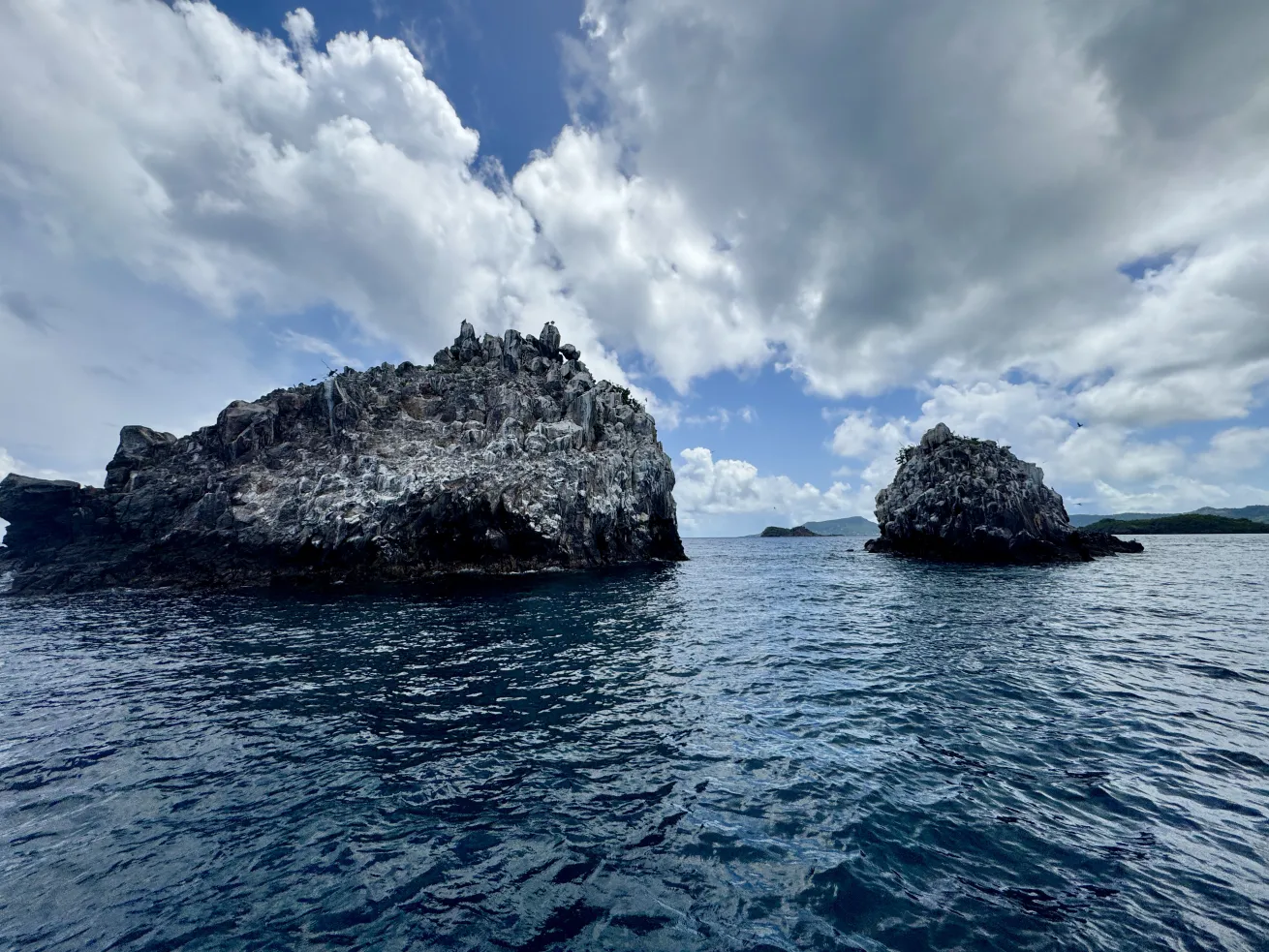 Rocky twin islets known as Sisters Rocks jut out of the ocean under a partly cloudy sky, with deep blue water in the foreground and distant green hills on the horizon.