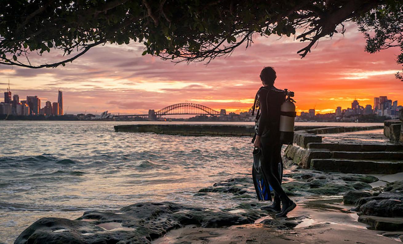 Male scuba diver stares at a cityscape across the water at sunset.