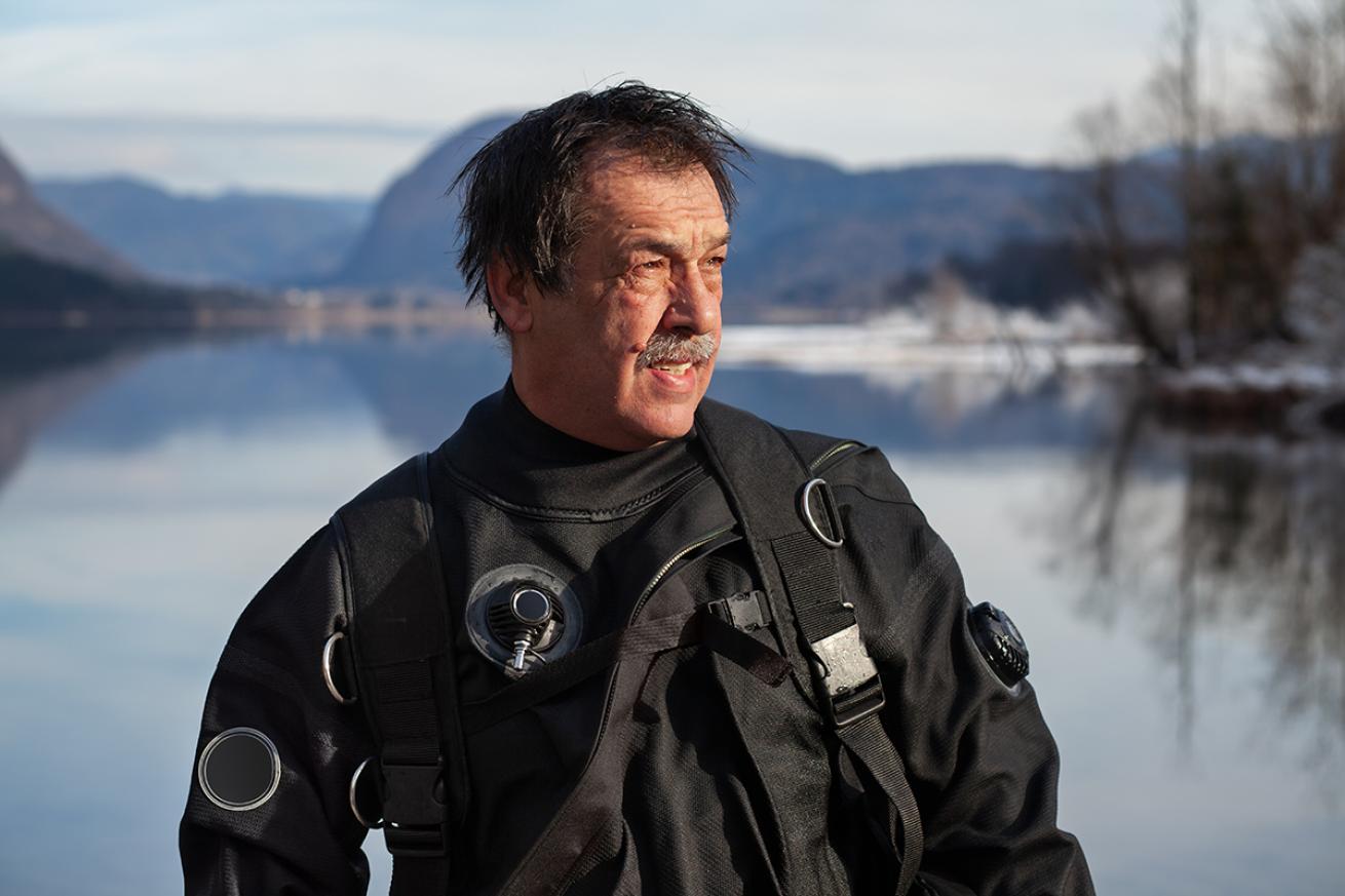 A mature diver in a drysuit stands in the foreground facing the camera with a lake and mountains in the background.