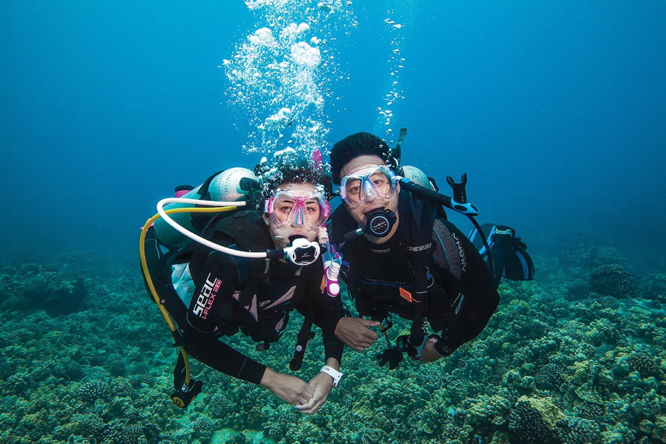 Two divers hover close to each other and a reef while looking straight at the camera.