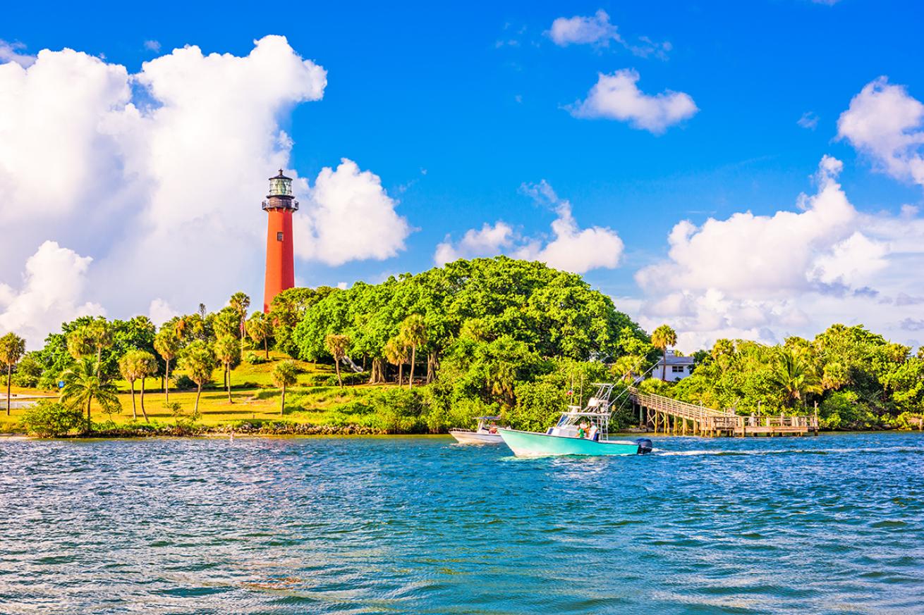A speedboat passes by a lighthouse overlooking an inlet in Jupiter, Florida on a sunny day.