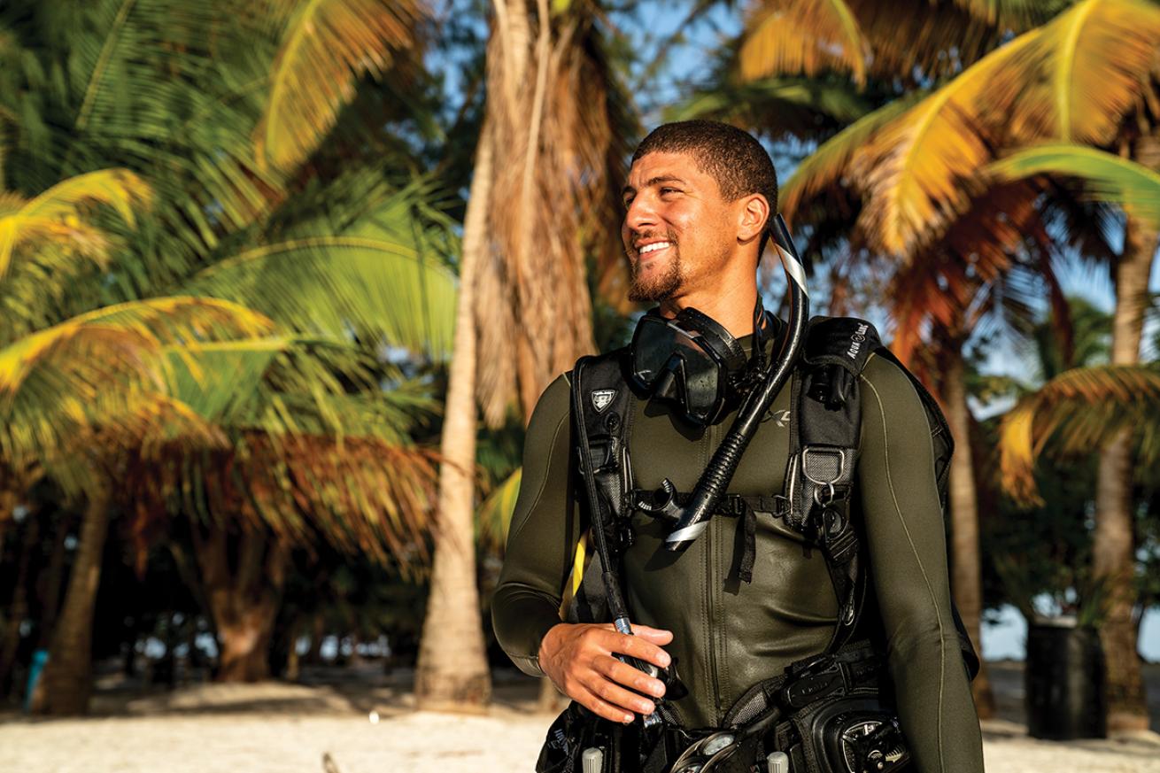 A diver stands on the beach with palm trees behind him.