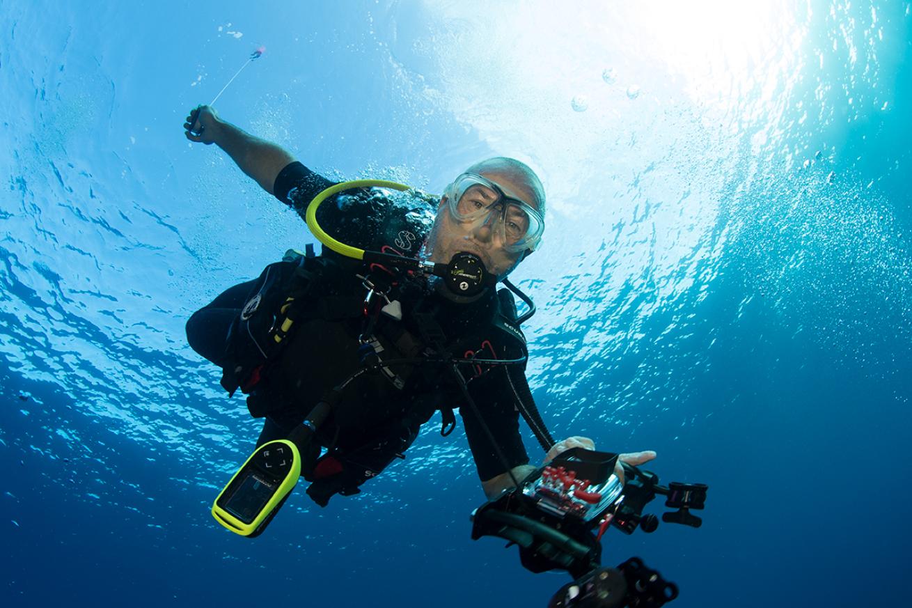 A diver holding photography equipment stares into camera in shallow water. 