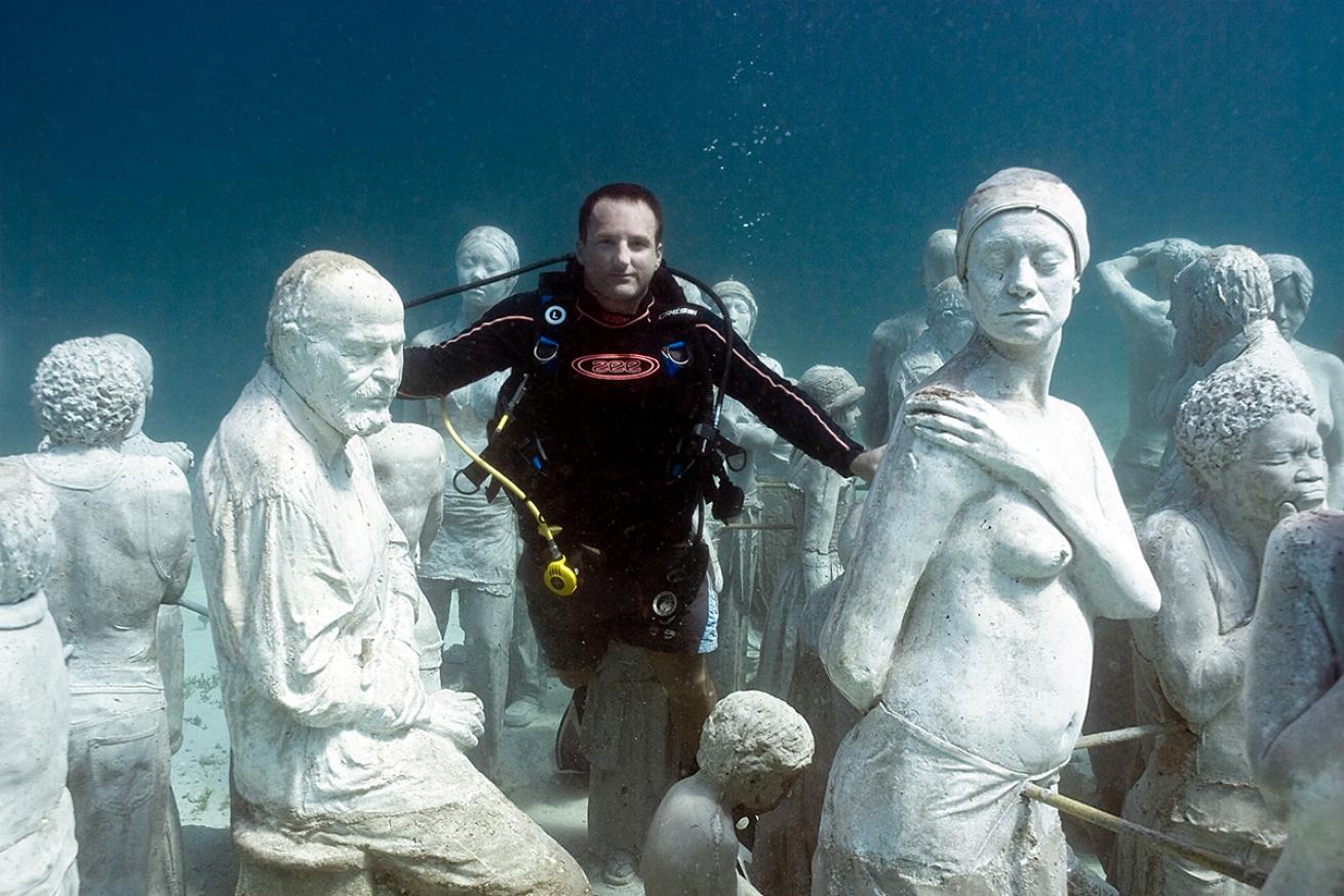 Jason de Caires Taylor hovers without a scuba regulator in his mouth among several underwater statues of his own creation
