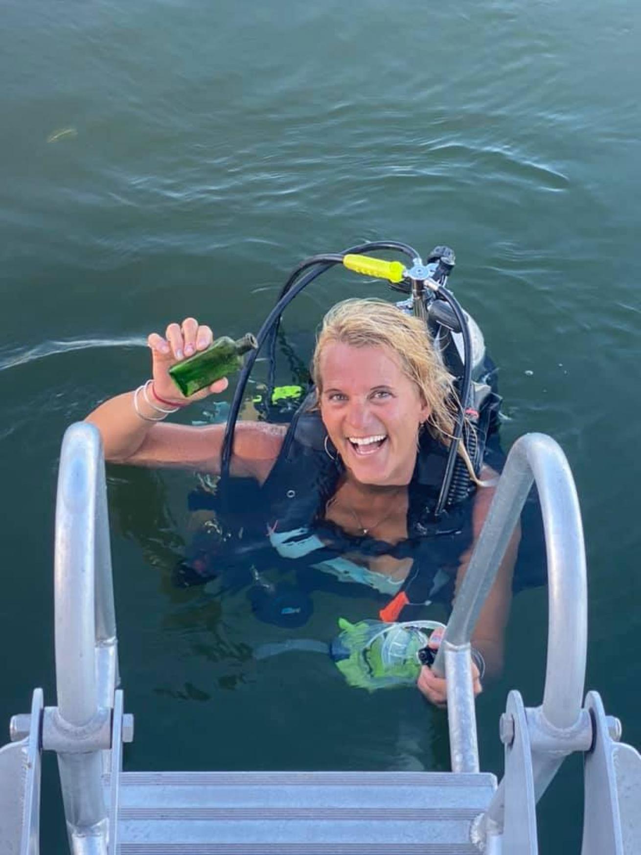 A female diver standing on boat ladder holds small bottle
