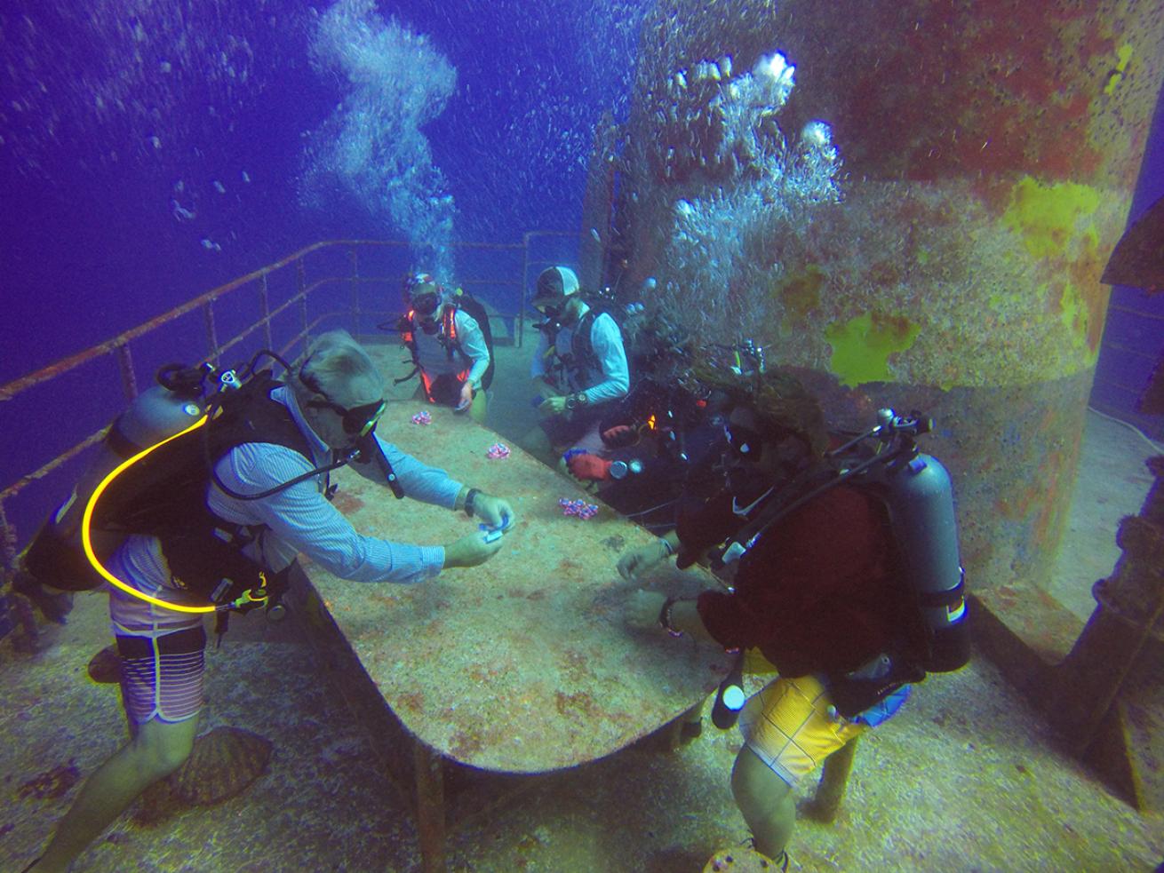 Five divers holding playing cards hover around a table on a shipwreck 