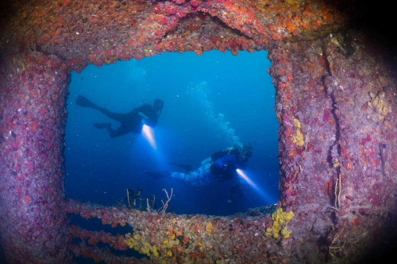 Two divers swimming by a shipwreck with flashlights