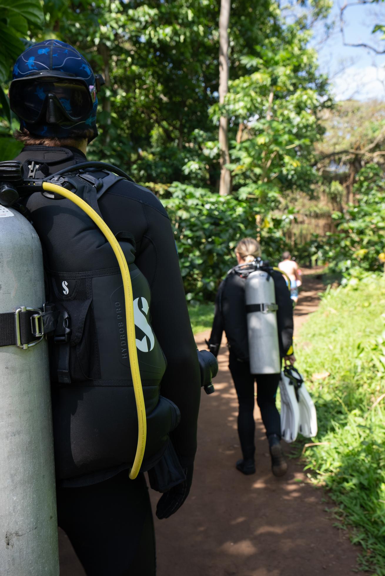 Scuba divers walk through a forest in Hawaii with scuba tanks on their backs to a dive site.