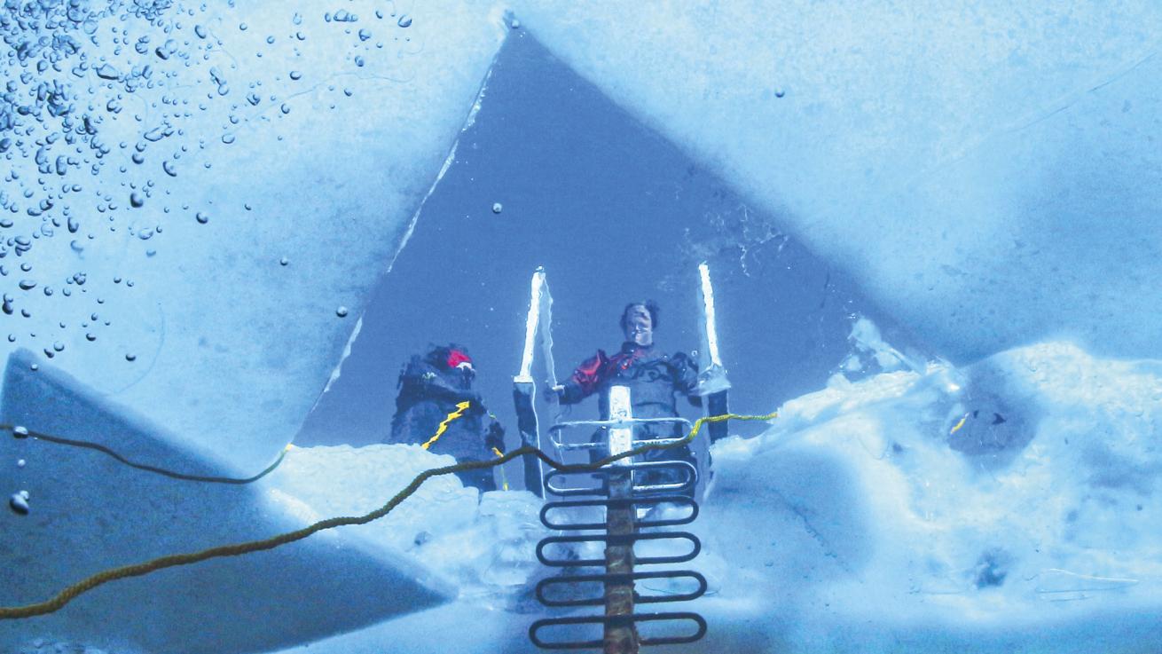 A drysuit-outfitted diver peers through a triangle-shaped entry point on the icy surface of White Star Quarry in Ohio.