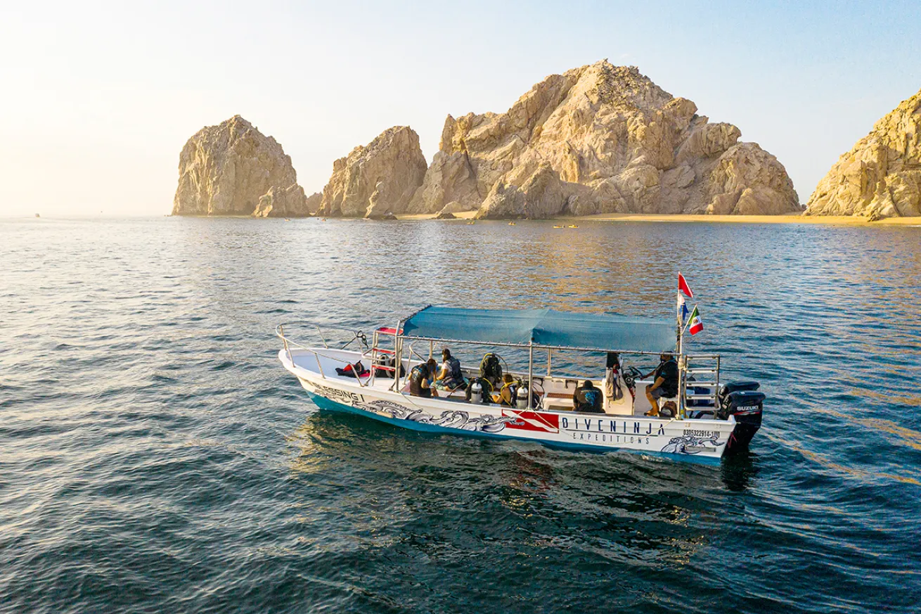 A small dive boat floats in blue water with large rocks emerging from the ocean in the background