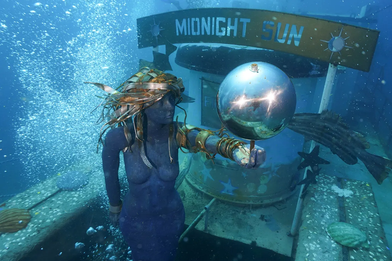 The statue of a mermaid holding a chrome ball sits on an algae wreck in front of a sign reading "Midnight Sun"