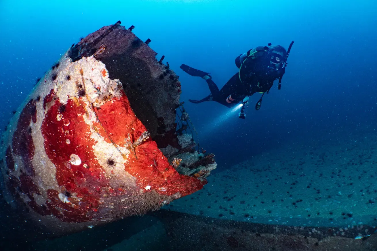 Underwater photograph showing a scuba diver exploring a large, rusted shipwreck covered in marine growth on ocean floor in Pensacola, Florida.