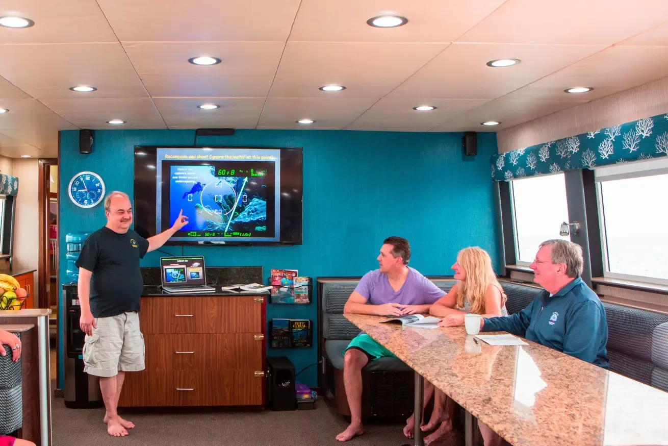 Photograph of a group dive briefing inside a scuba diving liveaboard, showing Aggressor staff standing and pointing at a screen displaying a navigation and ocean current map with data and directional arrows. Three seated individuals watch attentively around a granite table, with blue walls, a clock, and windows providing natural light in the background.