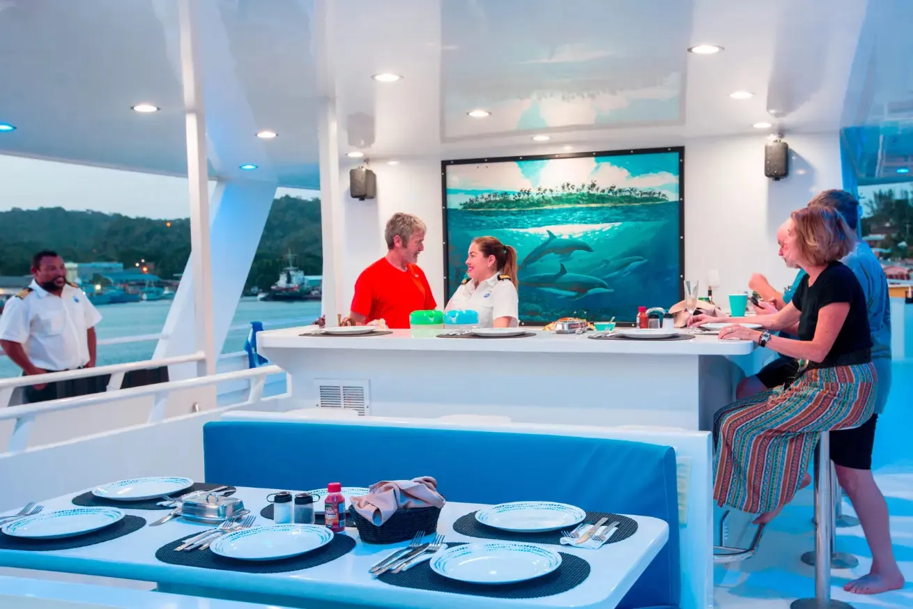 Photograph of a group of people socializing on a scuba diving liveaboard deck during sunset, with a table set for dining in the foreground. The scene includes a large screen displaying an underwater image of dolphins, adding a marine ambiance to the relaxed gathering.