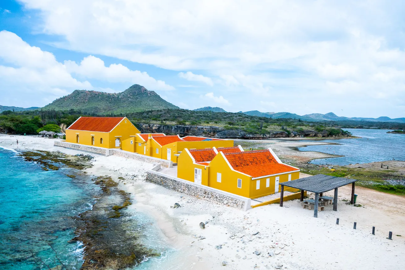 Photograph of bright yellow buildings in Bonaire with red roofs situated on a white sandy beach next to clear turquoise water and rocky shoreline.