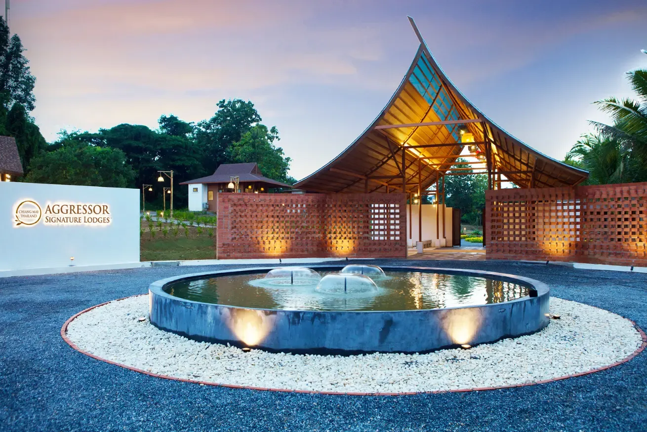 Photograph of a modern lodge entrance at dusk featuring a circular water fountain with three small jets in the foreground, surrounded by white stones and dark gravel.