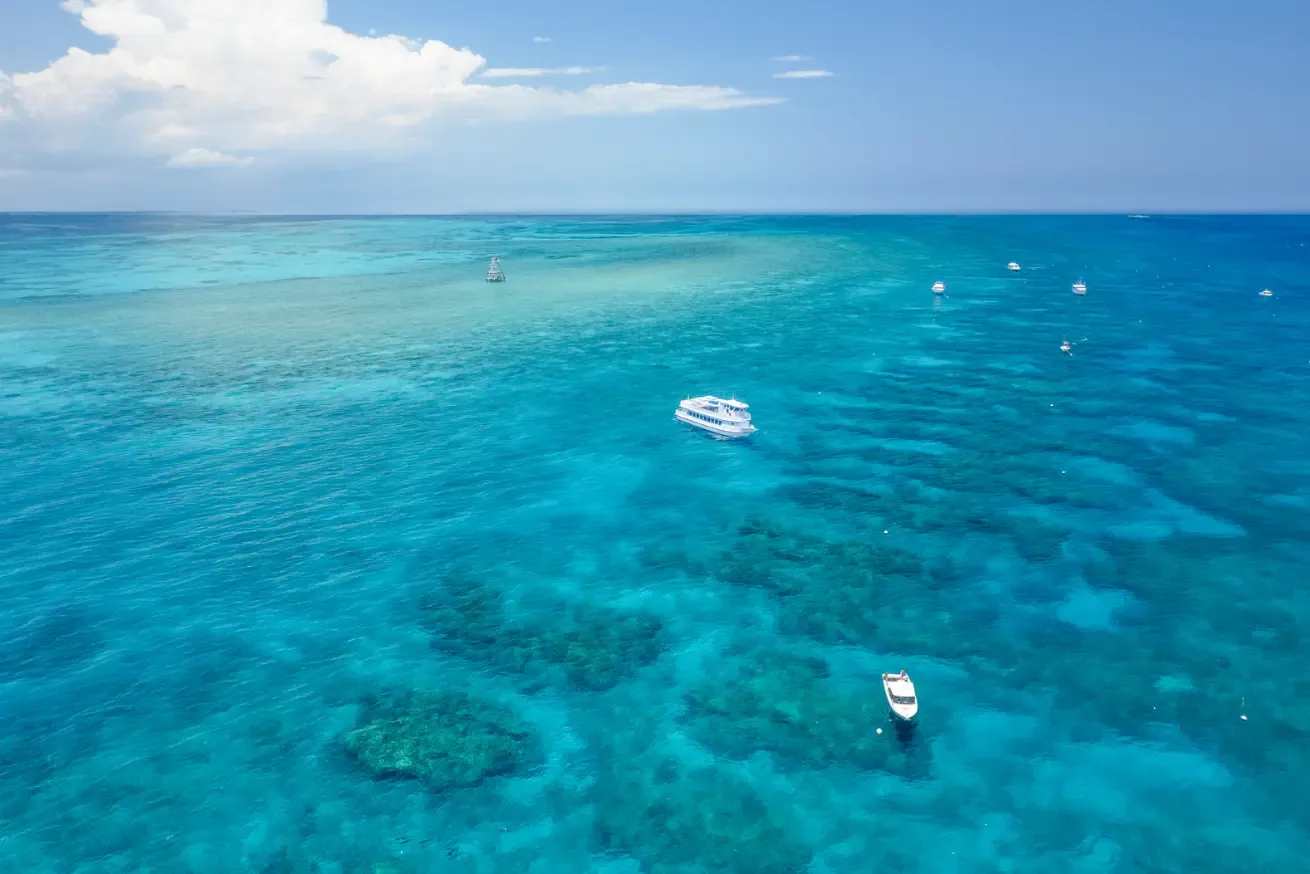 Photograph showing a vibrant blue ocean in Key Largo with several scuba diving boats scattered across clear, shallow waters, revealing coral formations beneath.