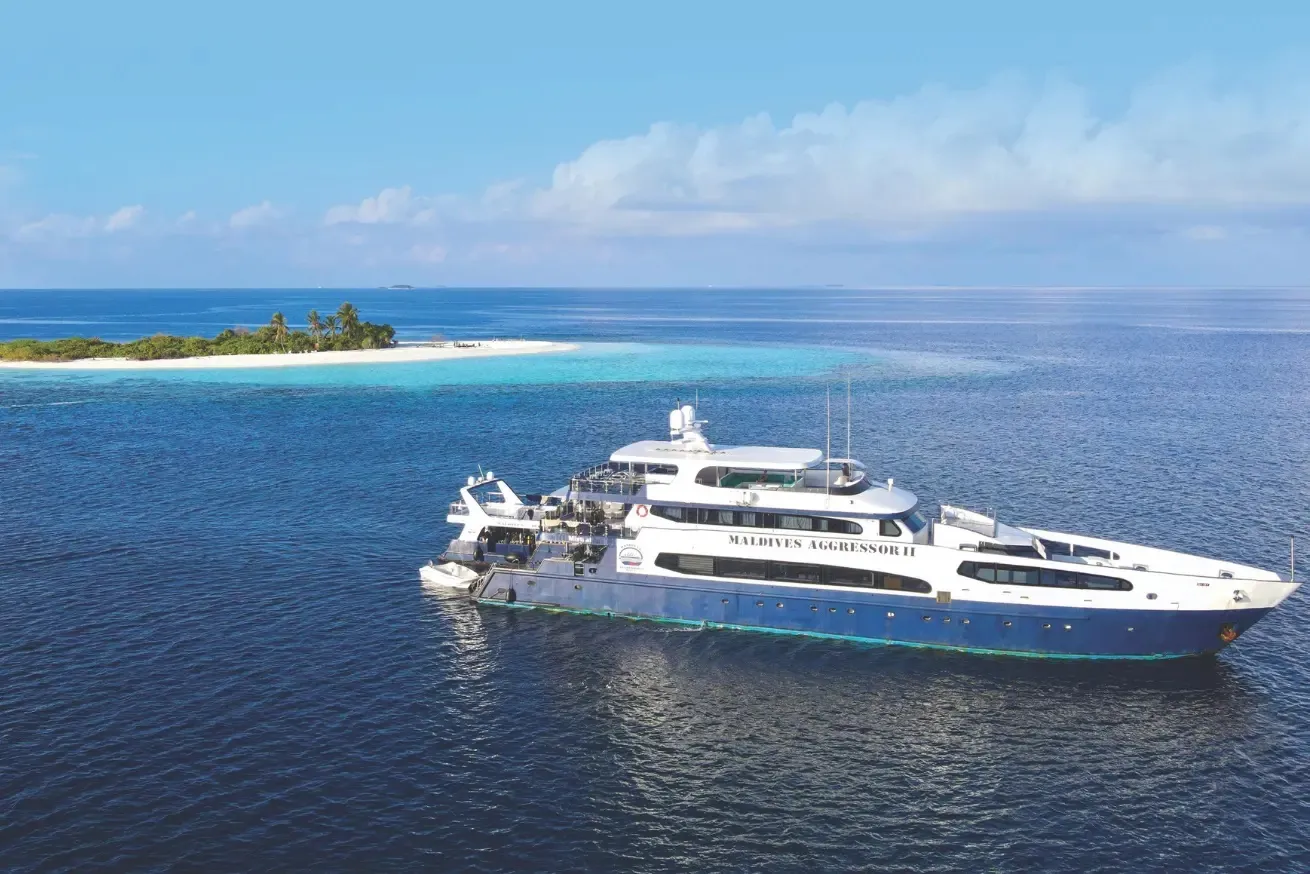 Photograph of a large white and blue yacht named "Maldives Aggressor II" sailing near a small tropical island with green vegetation and white sandy beach. The calm ocean water and partly cloudy sky create a serene maritime setting.