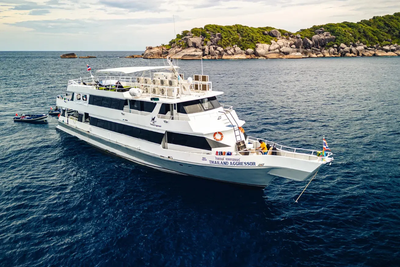 Photograph of a large white yacht named "Thailand Aggressor" anchored near a rocky, green island in calm blue waters.