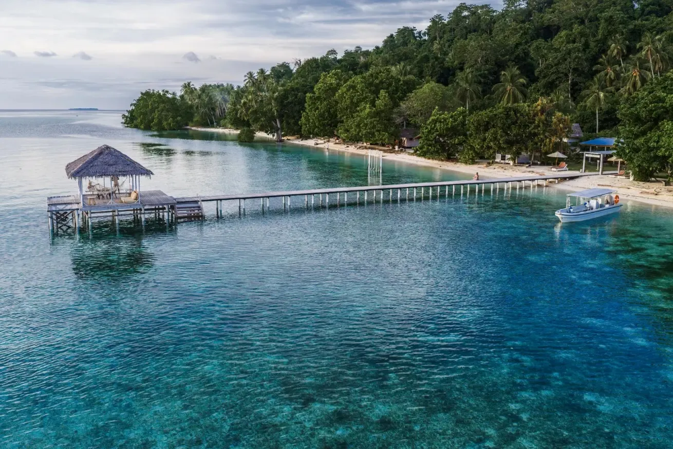 Aerial view of Reconnect Private Island Resort in Indonesia, featuring a wooden pier extending into clear turquoise water, a tropical beachfront lined with lush palm trees, and a small dive boat moored offshore.