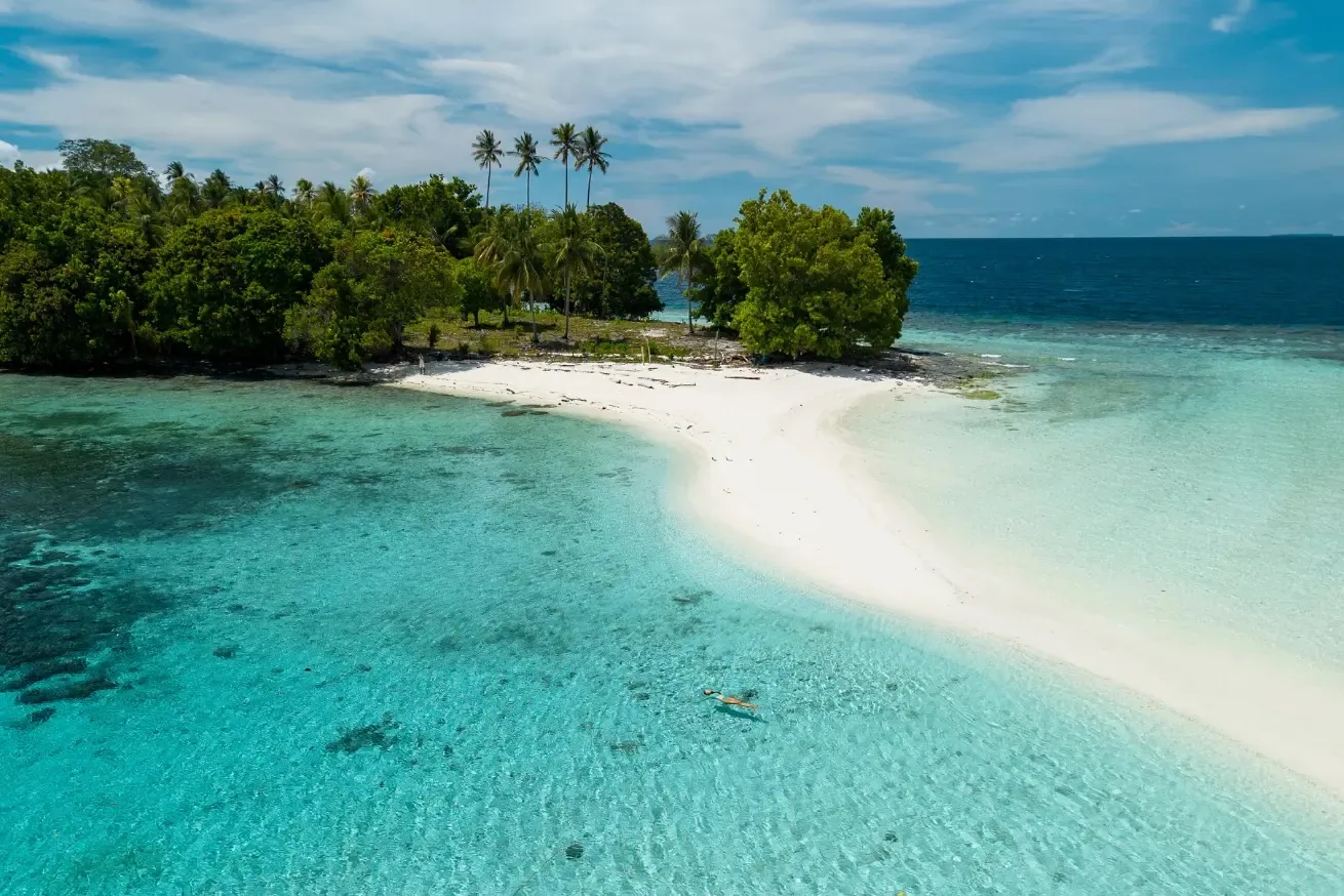 Aerial view of a white-sand beach and shallow turquoise lagoon at Reconnect Private Island Resort in Indonesia, with lush tropical forest, palm trees, and a lone swimmer floating in the clear water.