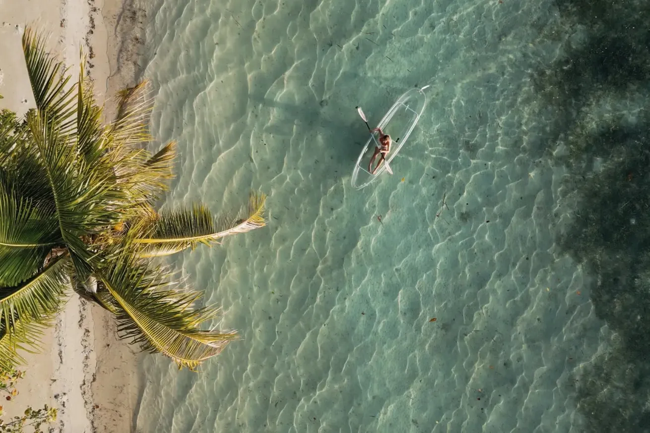 Aerial view of a guest kayaking in a transparent kayak through shallow turquoise water beside a white-sand beach lined with palm trees at Reconnect Private Island Resort in Indonesia.
