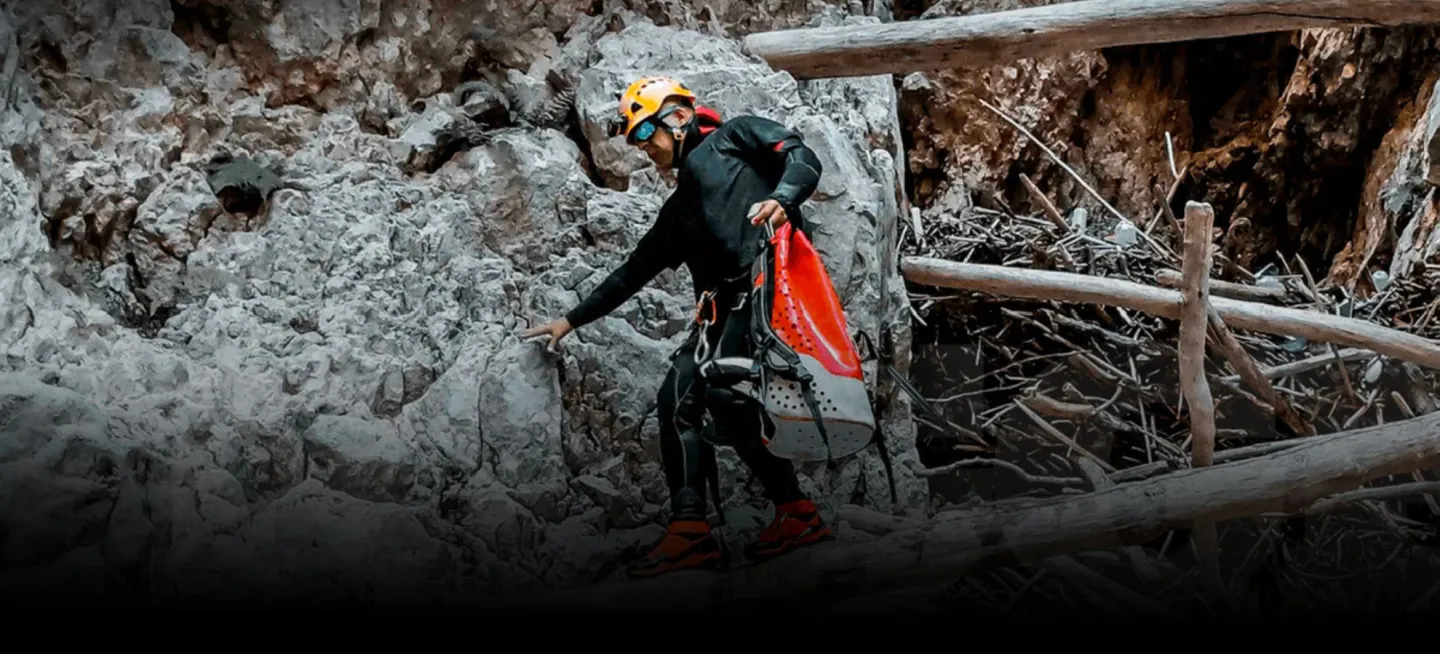A rugged outdoor scene shows a helmeted adventurer carefully navigating rocky terrain inside a cave or canyon, carrying gear.